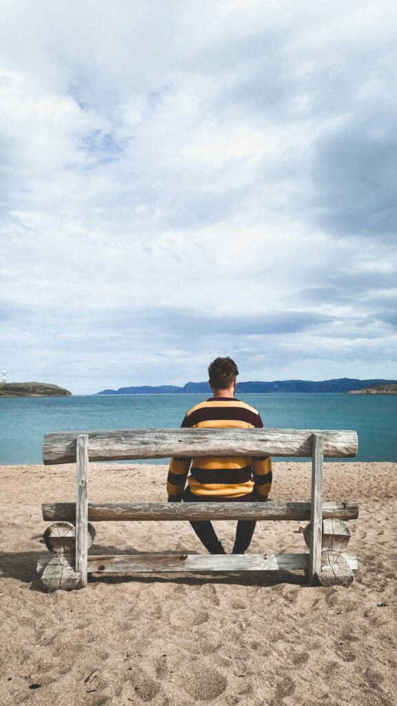 A man in a striped sweater sits on a wooden bench facing the sea under a cloudy sky, creating a tranquil scene.
